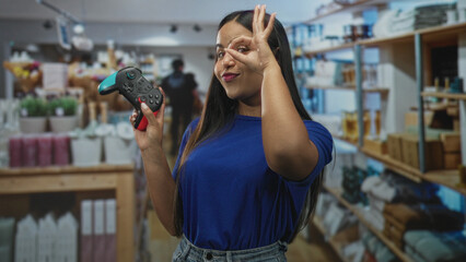 Woman holding gamepad with visible buttons making ok sign to eye inside building aisle with shelves, boxes and potted plants  playful fun social. © Krakenimages.com