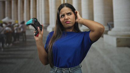 Young woman holds gamepad and shrugs palm up, showing hand and puzzled face in a columned building  confusion uncertainty. © Krakenimages.com