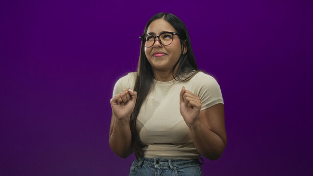 Young woman with glasses and hands raised, recoiling with a disgusted facial grimace, wearing beige tee and jeans in a purple studio; disgust avoidance.