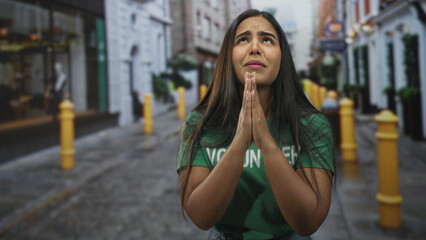 Fototapeta premium Hispanic volunteer woman with hands pressed together in praying gesture on a cobblestone street wearing a green volunteer shirt; pleading hope.
