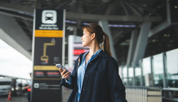 Young adult asian woman with travel luggage using smartphone app call taxi service transfer at international airport shuttle bus terminal