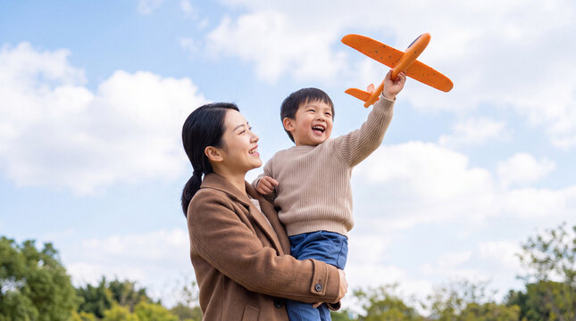 Asian woman holds smiling boy in her arms, child plays with orange toy airplane against a blue sky with fluffy clouds in a park setting