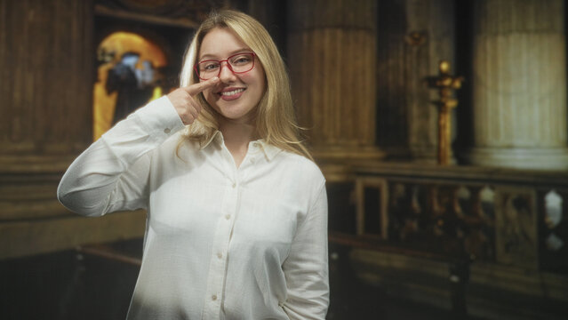 Woman wearing red glasses and white shirt, finger to nose gesture, smiling while standing by altar railing inside ornate building; playful joy.