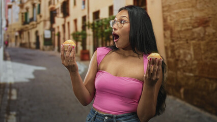 Obraz premium Woman brunette wearing a pink top and jeans holds muffins in both hands on a street, mouth open and looking upward; joyful wonder.