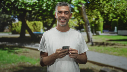Obraz premium Man smiling and looking down while holding smartphone with both hands and tapping screen in a city park pathway under trees; contentment connection.