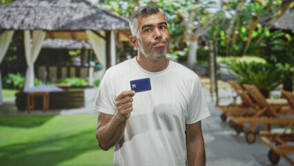 Fototapeta premium Middle age man with grey hair holding creditcard, looking up with puzzled face in resort building; financial worry uncertainty.