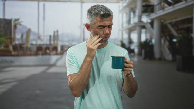 Man holding teal mug presses cheek with wince and closed eyes for toothache inside a bright building concourse; discomfort morning routine.