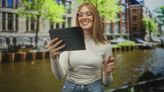 Woman redhead with glasses holding tablet and gesturing with hand on a canal street by water and buildings, wearing white top and jeans; curiosity.