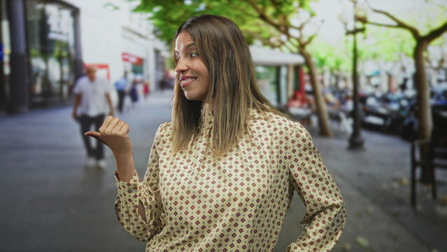 Woman pointing thumb to the side with visible hand gesture on a street, smiling with casual posture; confidence playfulness.