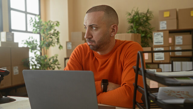 Man in orange sweater checks laptop and watch, hand on chin, barcode scanner on desk in building; small business frustration.