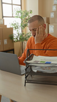Man hand on face over laptop amid stacked cardboard boxes and desk papers in parcel office building, facepalm and paused typing; small business stress.