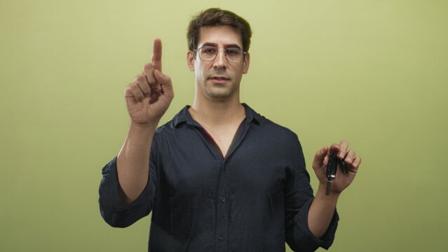 Man wearing glasses points finger while holding car keys against a green studio backdrop with neutral expression; instructional choice.