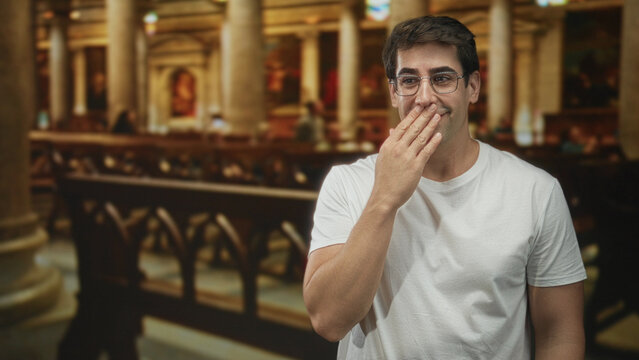 Man in white t shirt with glasses raises hand to mouth, gesturing while speaking inside ornate churchlike building with pews and columns; warm amusement.