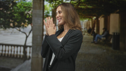 Fototapeta premium Woman claps hands on tree lined street beside stone columns, smiling in black pinstripe blazer and white top; joy gratitude.