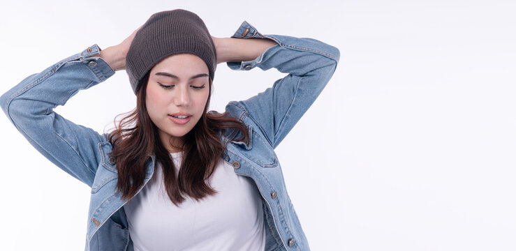 Beautiful young Asian woman in casual blue denim jacket and grey beanie hat posing with hands behind head and looking down on white background with copy space. Relaxed winter fashion concept.