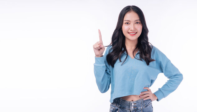 Beautiful smiling young Asian woman in casual blue sweater pointing finger up at copy space on white background. Happy female student presenting idea or recommending product concept.
