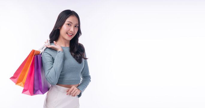 Portrait of a beautiful smiling young Asian woman carrying colorful shopping bags over her shoulder, looking at camera on white background with copy space. Happy female shopper retail concept.