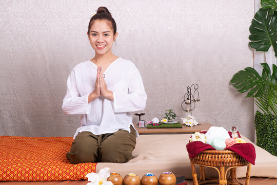 Beautiful young Asian Thai massage therapist in white uniform smiling and making wai greeting gesture while sitting on massage bed in spa salon. Welcoming wellness and relaxation concept.