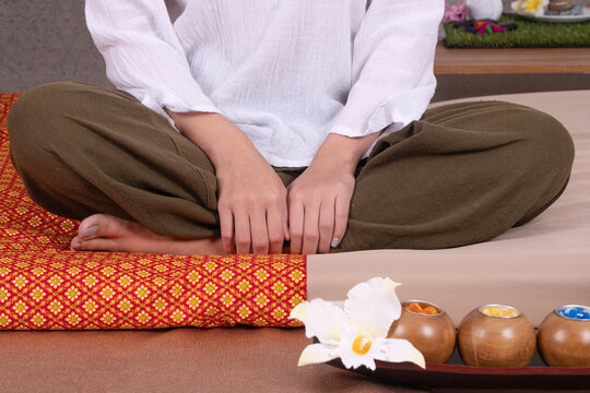 Close up of massage therapist in uniform sitting cross legged on bed with traditional thai mat orchid flower and aromatic candles. Relaxing spa wellness and alternative medicine concept.
