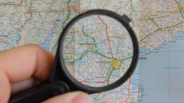 Man's hand holds a magnifying lens to look at a classic paper map of northern Italy