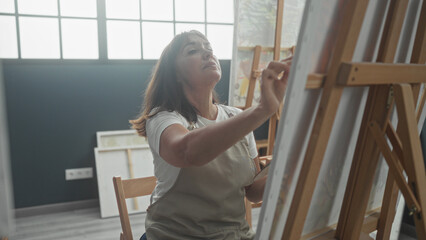 Woman painter wearing apron, holding palette and brush, painting canvas with hand at easel in studio  creative concentration. © Krakenimages.com