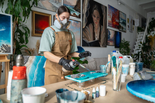 Focused craftswoman creating fluid art painting in gallery workshop using gas torch and epoxy resin, woman artisan holding blowtorch and heats up surface with liquid resin on canvas to remove bubbles.