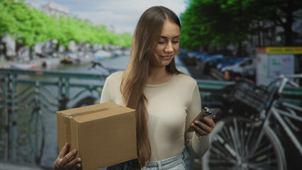 Naklejka premium Woman holding box and checking phone on a street bridge, smiling with hands gripping package; contentment routine.