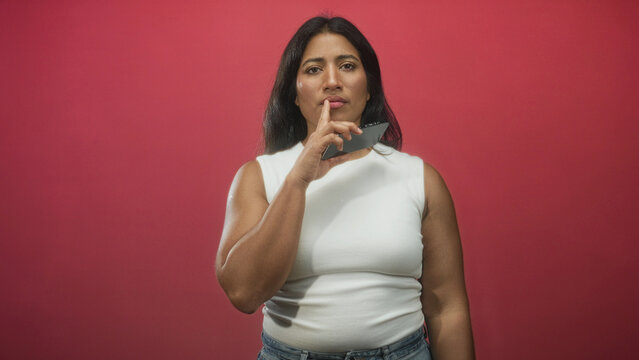 Woman holding smartphone with finger to lips for hush in studio, wearing white sleeveless top and blue jeans; thoughtful curiosity.