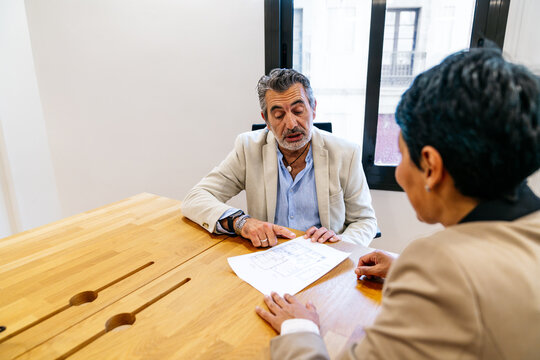 Two businesspeople reviewing an architectural blueprint on a wooden table, collaborating and discussing a project in a modern office