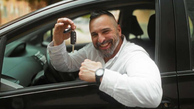 Middle age man holding car keys with raised hand and fist pump while leaning from a car window on street, smiling broadly showing teeth, white shirt and wristwatch visible; joy success.