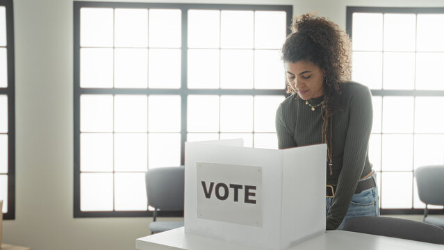 Woman marking a ballot at a voting booth inside a public building, leaning over the privacy screen to cast a vote; civic determination.