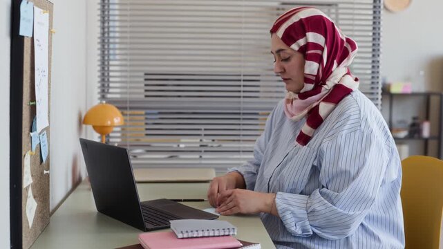 Young adult Middle Eastern Muslim woman wearing hijab writing notes, pinning sticky note to corkboard, and working on laptop at desk. Footage ideal for corporate diversity and remote work campaigns