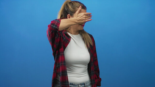 Woman covers eyes with hand and shields face while standing against a blue studio backdrop, wearing plaid shirt and white top; shyness privacy.