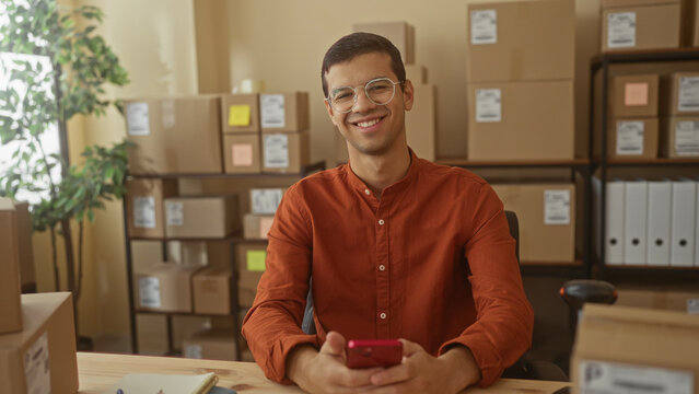 Man holding smartphone and smiling amid stacked shipping boxes at a small business packing desk inside building; entrepreneurial confidence.