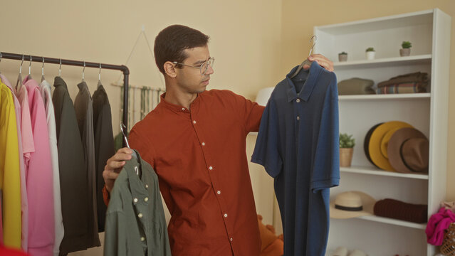 Young hispanic man comparing a green button shirt and a blue polo on hangers among clothing racks and hats inside a retail building; decision.