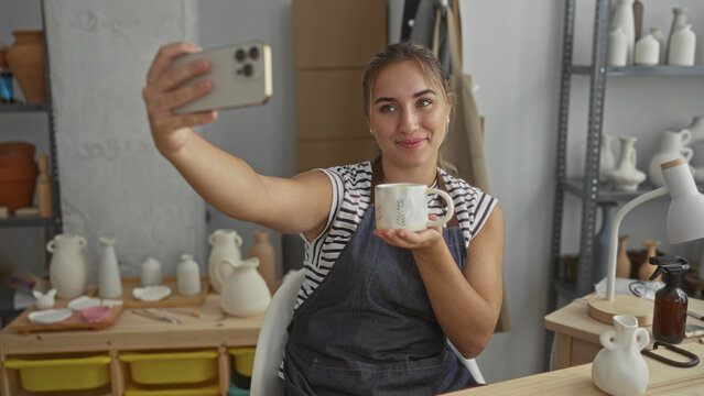 Woman taking selfie with smartphone while holding painted ceramic mug and smiling, wearing apron amid pottery shelves in studio; creative joy.