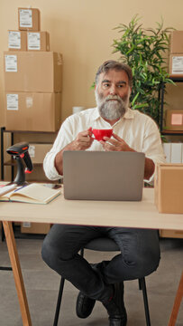 Man with gray beard holding red mug with both hands at a laptop on a desk surrounded by shipping boxes and a barcode scanner in building; calm focus.