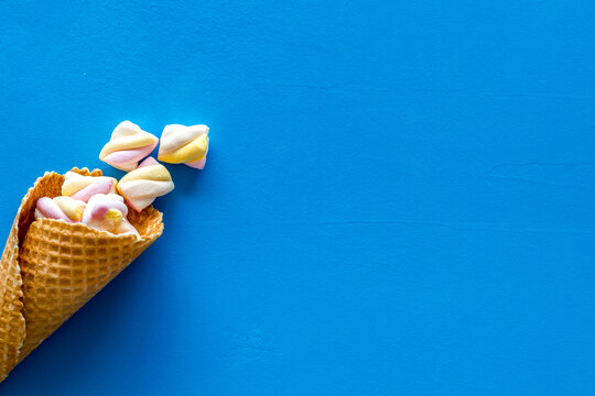Sweet desserts. Marshmallows in ice cream waffle cone on blue background top view space for text