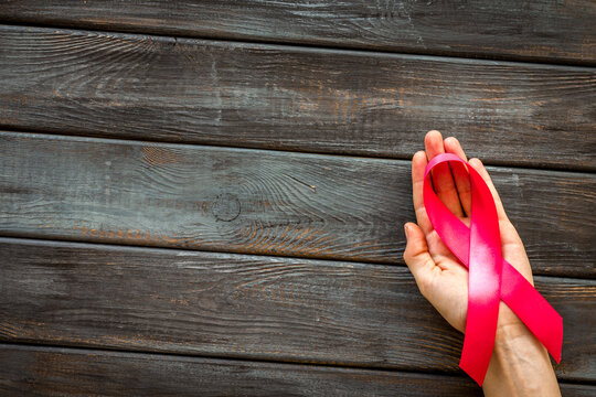 Pink ribbon in hands as symbol of breast cancer awareness on dark wooden background top view copy space