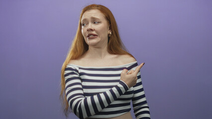 Obraz premium Redhead woman points both index fingers with crossed arms exposing bare shoulder while wearing striped top in studio; confusion doubt.