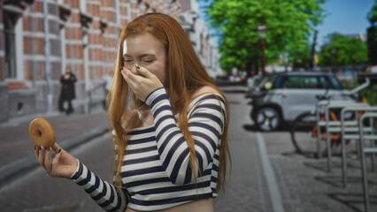 Fototapeta premium Redhead woman holding donut with hand to mouth laughing on urban street, striped top visible; carefree joy.