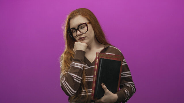 Redhead woman holding a large leather book with hand under chin wearing striped sweater and eyeglasses in studio; contemplation.