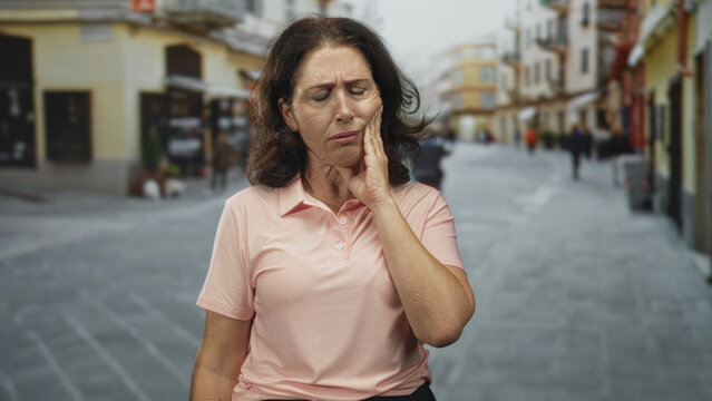 Middle age woman with hand on cheek on a pedestrian street, facial wince and closed eyes suggest tooth pain; discomfort toothache.