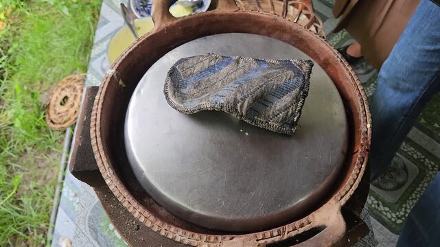 Hand covering a traditional clay pan with a metal lid using a cloth to cook Vietnamese mini savory pancakes.