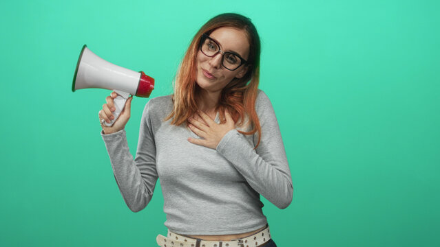 Woman redhead speaking into megaphone, hand on chest in studio with teal backdrop and gray sweater; enthusiasm empowerment.