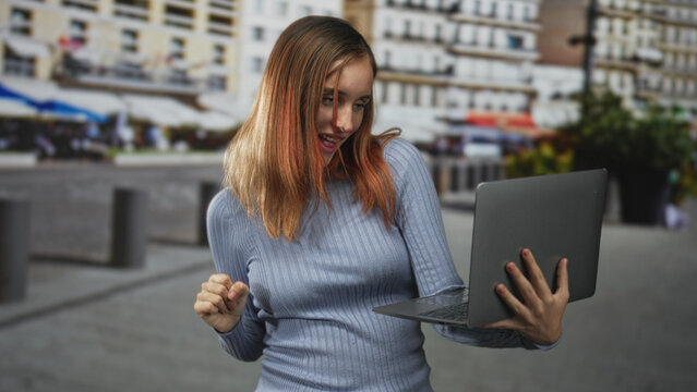 Young woman holding a laptop and pumping her fist while smiling on a street plaza amid cafe buildings; joy success.