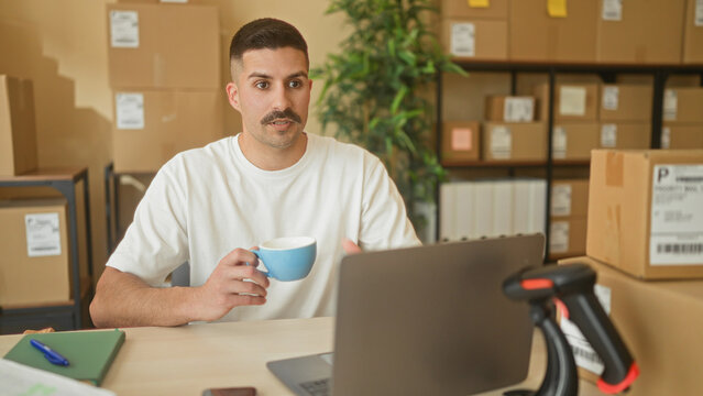 Man holding a mug while working on a laptop and checking packages in a small business building; quiet concentration.