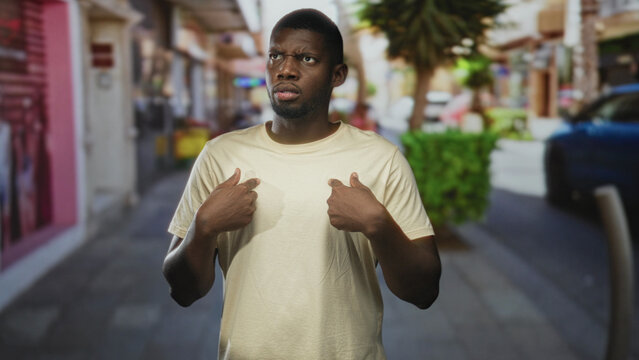 Young man pointing to his chest with both hands on a busy street, beige tshirt and puzzled expression, looking sideways; confusion self doubt.