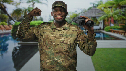 Naklejka premium Man soldier in camouflage holding binoculars with fist raised beside a poolside building, smiling and surveying the area; pride duty vigilance.