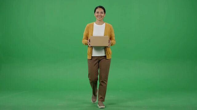 Smiling young woman walking towards camera holding a cardboard box on a green screen.